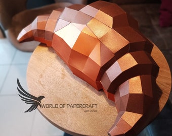 May include: A copper-colored papercraft croissant, made of folded paper, sits on a wooden table. The croissant is a realistic representation of a baked pastry, with a detailed, textured surface.