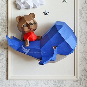 Puede incluir: Un modelo de papel de una ballena azul con un oso marrón sentado encima. La ballena y el oso están sobre un fondo blanco con nubes de papel blanco y estrellas plateadas.