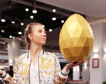 May include: Three large geometric papercraft eggs in gold, black, and white. The eggs are displayed on green cube pedestals in a retail setting. The text "WORLD OF PAPERCRAFT ARTSTORE" is visible in the lower right corner of the image.