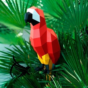 Puede incluir: Un loro de papel rojo, amarillo y negro posado sobre un follaje verde. El loro está hecho de papel doblado y tiene un diseño geométrico detallado.