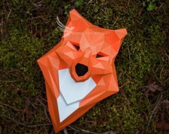 Può includere: Maschera di volpe in cartapesta arancione e bianca con un design geometrico. La maschera è sdraiata su un letto di muschio verde.