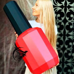 Puede incluir: Una mujer sostiene una botella grande de esmalte de uñas rojo y negro. La botella está hecha de papel y tiene un diseño geométrico.