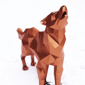 Puede incluir: Una escultura de lobo geom&eacute;trico de color cobre con un dise&ntilde;o estilizado. El lobo est&aacute; de pie sobre una superficie blanca y est&aacute; aullando con la cabeza echada hacia atr&aacute;s.