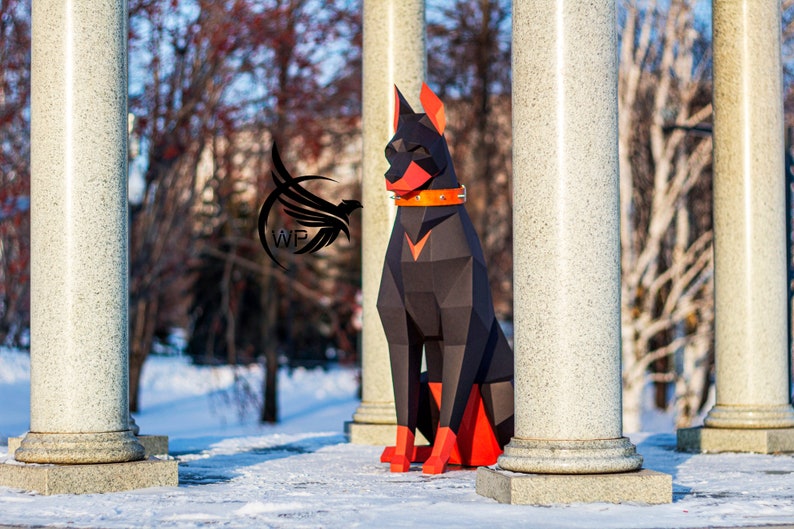 Peut inclure: Sculpture en papier d'un Dobermann Pinscher noir et orange assis devant des colonnes blanches. Le chien porte un collier en cuir marron.