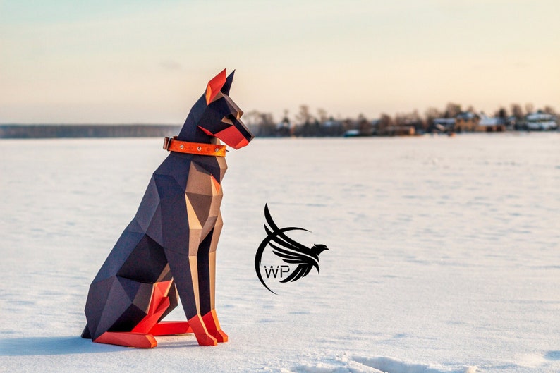 Peut inclure: Un chien Doberman Pinscher en papiercraft noir et orange assis sur une surface enneig&eacute;e. Le chien porte un collier orange avec une boucle en m&eacute;tal.