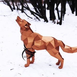 Puede incluir: Una escultura de lobo de origami dorada se alza sobre un suelo nevado con un bosque en el fondo. El lobo a&uacute;lla con la cabeza echada hacia atr&aacute;s y la boca abierta.