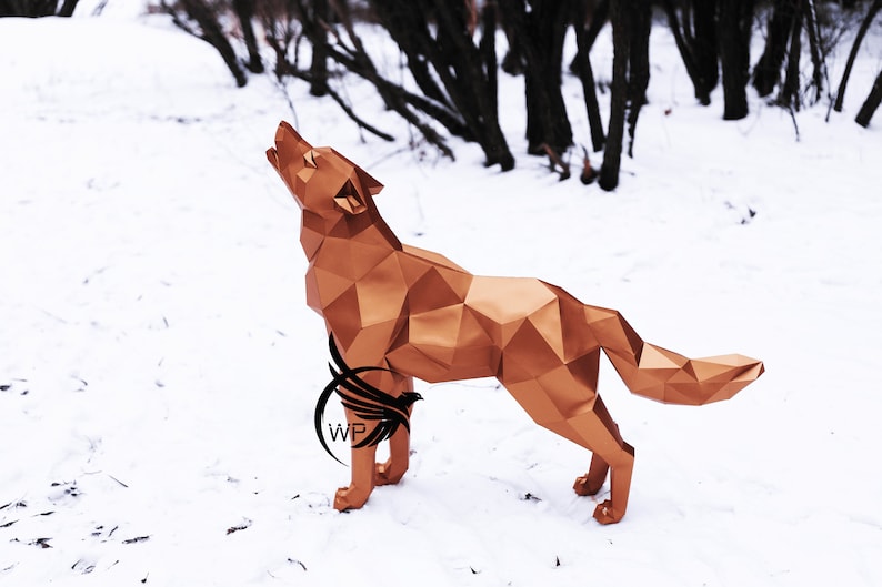 Puede incluir: Una escultura de lobo de origami dorada se alza sobre un suelo nevado con un bosque en el fondo. El lobo a&uacute;lla con la cabeza echada hacia atr&aacute;s y la boca abierta.