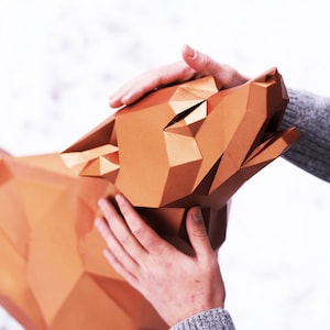 Puede incluir: Una mano toca suavemente una gran escultura de lobo geom&eacute;trica de color cobre. La escultura est&aacute; hecha de papel y tiene un dise&ntilde;o facetado detallado.