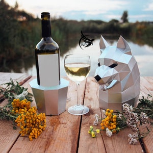 Puede incluir: Un porta botellas de vino plateado y una copa de vino blanco sobre una mesa de madera con una escultura de lobo de origami plateada. La mesa está al aire libre cerca de un cuerpo de agua.