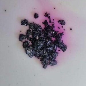 Op de afbeelding: Een cluster van kleine, onregelmatige, donkerpaarse tot zwarte kristallijne fragmenten met een metaalachtige glans. De stukjes liggen in een losse hoop en vormen een scherp contrast met de lichte ondergrond met een vage roze vlek.