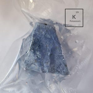 Può includere: Primo piano di un pezzo di metallo di potassio, un metallo alcalino morbido e bianco argenteo, in un sacchetto di plastica trasparente. Il metallo è reattivo e si opacizza rapidamente all'aria. L'immagine include anche il simbolo chimico del potassio, "K", e il suo numero atomico, "19".