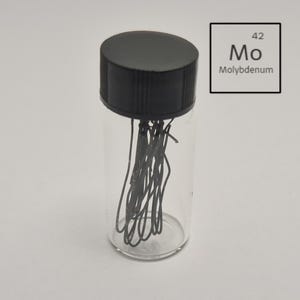 May include: A clear glass vial with a black cap, containing several strands of dark grey material. A white square with the number 42, the symbol Mo, and the word Molybdenum is in the background.
