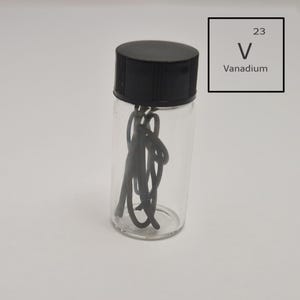 Peut inclure: Un flacon en verre transparent avec un bouchon noir contenant un métal gris foncé enroulé. Un carré blanc avec le texte "23 V Vanadium" est en arrière-plan. Le flacon est sur une surface blanche.
