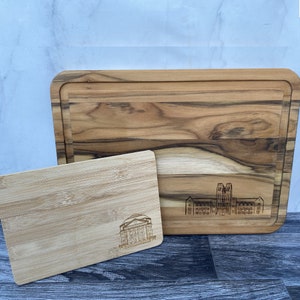 May include: Two wooden cutting boards, one larger and one smaller. The larger board is made of teak wood and has a natural wood finish. The smaller board is made of bamboo and has a light brown finish. Both boards feature laser-engraved images of buildings.