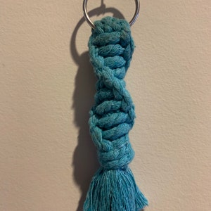 May include: A teal blue macrame keychain with a silver key ring. The keychain is made of twisted rope with a tassel at the bottom.