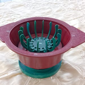 May include: A red plastic flower pot with a green plastic flower holder inside. The flower holder has multiple prongs that are designed to hold flowers in place.
