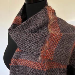 May include: A woven scarf featuring a dark charcoal gray base with horizontal stripes in a vibrant burnt orange. The fabric has a textured, chunky knit appearance with a frayed, fuzzy orange trim along the edge of the wrap-style garment.