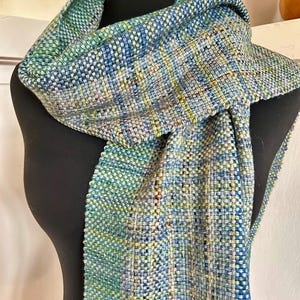 May include: A handwoven scarf featuring a variegated pattern of blue, teal, green, and yellow threads. The textile displays a tight, consistent weave with a visible grid-like texture throughout, creating a multi-colored appearance across the fabric.