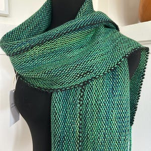 May include: A handwoven scarf featuring a variegated blend of emerald, forest, and lime green yarns with distinct black vertical stripes. The textile displays a dense, textured weave pattern and includes a small white paper tag attached to the side.