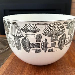 May include: White ceramic bowl with a black mushroom pattern. The bowl has a rounded shape and a black rim. The mushroom illustrations vary in size and style, creating a whimsical design. The bowl is sitting on a wooden surface.