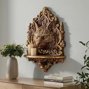 May include: Ornate, golden cat-faced wall shelf with a gothic design. The cat's face is intricately detailed, and the shelf includes a small ledge for displaying items. Two white candles sit on the shelf. A unique home decor piece.