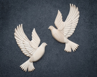 May include: Two white wooden doves with detailed carving, flying in opposite directions against a dark gray background.