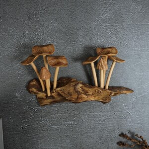 May include: A decorative wall sculpture featuring two clusters of carved wooden mushrooms attached to a piece of textured driftwood. The mushrooms have light brown, ribbed caps and slender stems, while the base is a dark, weathered piece of wood.