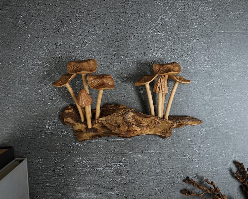 May include: A decorative wall sculpture featuring two clusters of carved wooden mushrooms attached to a piece of textured driftwood. The mushrooms have light brown, ribbed caps and slender stems, while the base is a dark, weathered piece of wood.