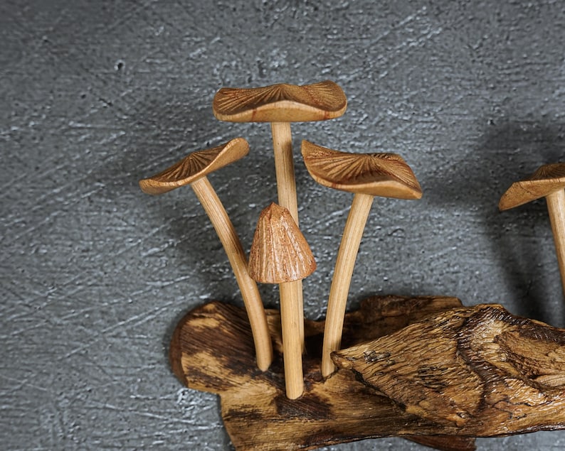 May include: A decorative sculpture featuring several carved wooden mushrooms with light brown stems and textured caps, attached to a piece of dark, rough-hewn natural wood. The mushrooms vary in height and cap shape, displaying detailed wood grain.
