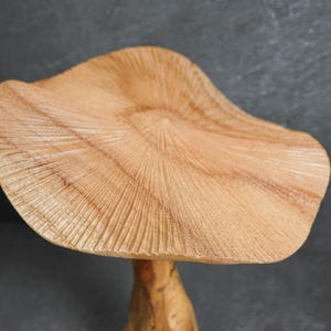 May include: A light brown wooden mushroom sculpture featuring a wide, wavy cap with detailed, radiating carved lines across the surface. The texture of the wood grain is visible throughout the cap, which sits atop a smooth, cylindrical stem.