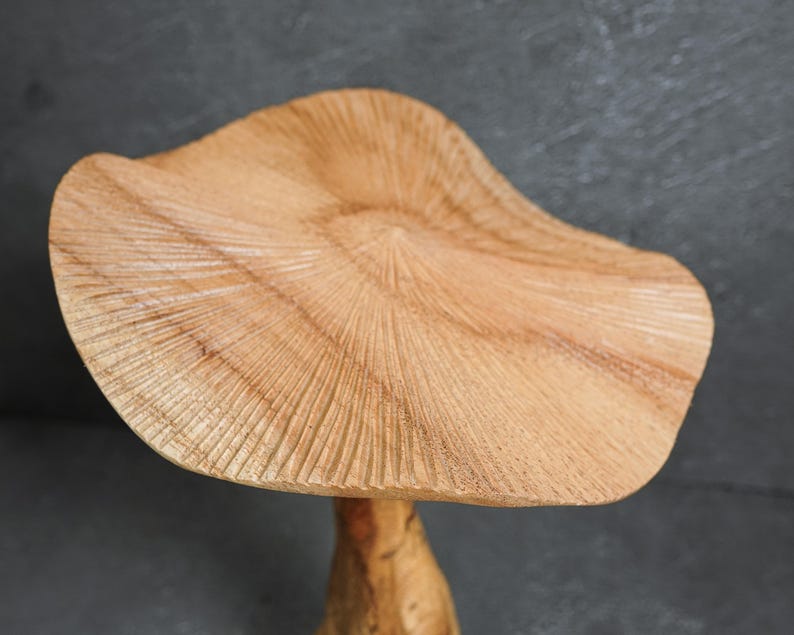 May include: A light brown wooden mushroom sculpture featuring a wide, wavy cap with detailed, radiating carved lines across the surface. The texture of the wood grain is visible throughout the cap, which sits atop a smooth, cylindrical stem.