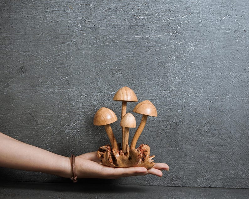 Pu&ograve; includere: Una scultura decorativa composta da quattro funghi in legno marrone chiaro con cappelli lisci e arrotondati e steli sottili, tutti fissati su una base di legno naturale nodoso e strutturato che funge da supporto per il gruppo di funghi.