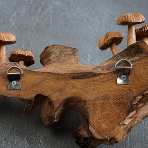 May include: A natural wood piece featuring several carved wooden mushrooms attached to the surface. Two silver-colored metal D-ring hangers are screwed into the back of the wood, allowing for wall mounting. The wood grain is visible throughout.