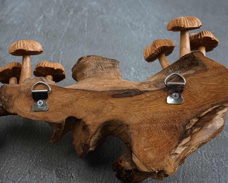 May include: A natural wood piece featuring several carved wooden mushrooms attached to the surface. Two silver-colored metal D-ring hangers are screwed into the back of the wood, allowing for wall mounting. The wood grain is visible throughout.
