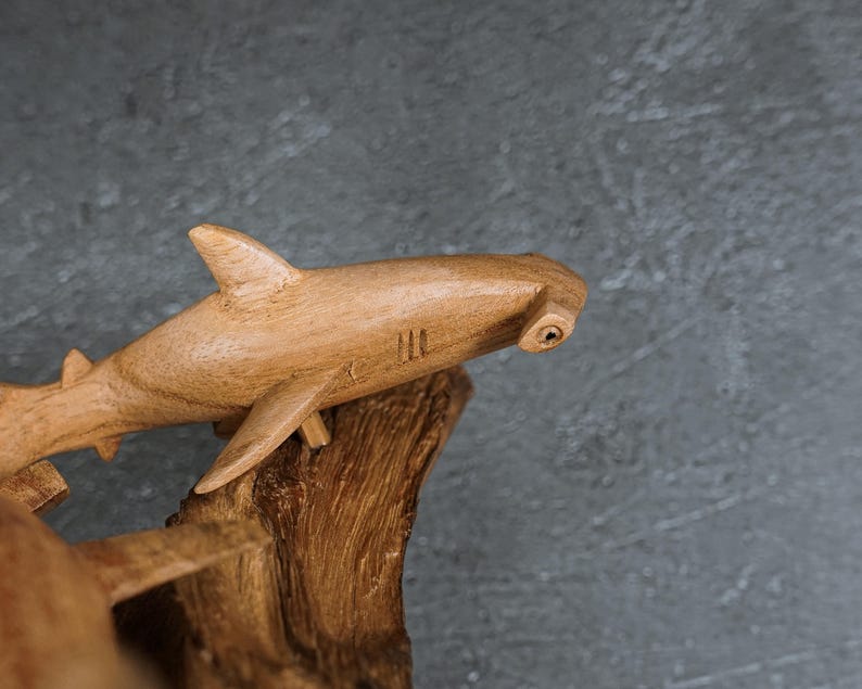 May include: A wooden hammerhead shark sculpture with visible grain and a natural finish. The shark is mounted on a piece of driftwood, showcasing the craftsmanship and detail of the carving. The sculpture has fins and gills.
