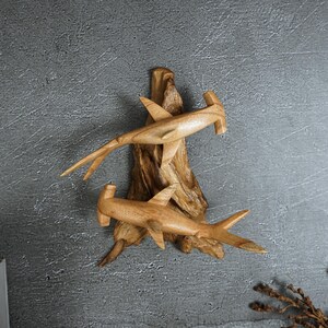 May include: Wooden sculpture featuring two hammerhead sharks carved from light-colored wood. The sharks are mounted on a piece of driftwood, creating a natural and artistic display. The sculpture is a decorative item.