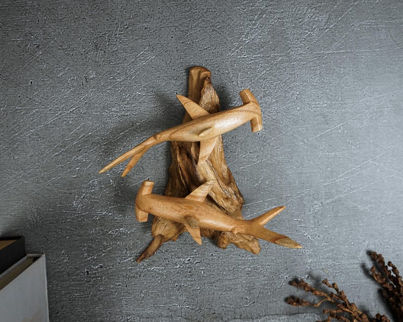 May include: Wooden sculpture featuring two hammerhead sharks carved from light-colored wood. The sharks are mounted on a piece of driftwood, creating a natural and artistic display. The sculpture is a decorative item.