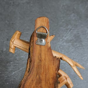 May include: Close-up of a handcrafted wooden wall decor piece. The natural wood grain is visible, with a metal hanger attached. The design incorporates a hammer and antler-like elements, showcasing a rustic aesthetic. The piece is a warm brown color.