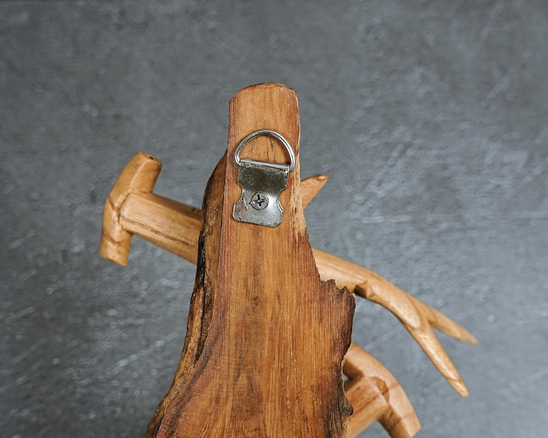 May include: Close-up of a handcrafted wooden wall decor piece. The natural wood grain is visible, with a metal hanger attached. The design incorporates a hammer and antler-like elements, showcasing a rustic aesthetic. The piece is a warm brown color.