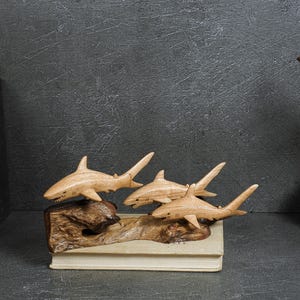 May include: A wooden sculpture featuring three carved sharks attached to a piece of driftwood. The sharks are light brown with visible wood grain, and the base is a dark, textured piece of natural wood resting on a rectangular book.