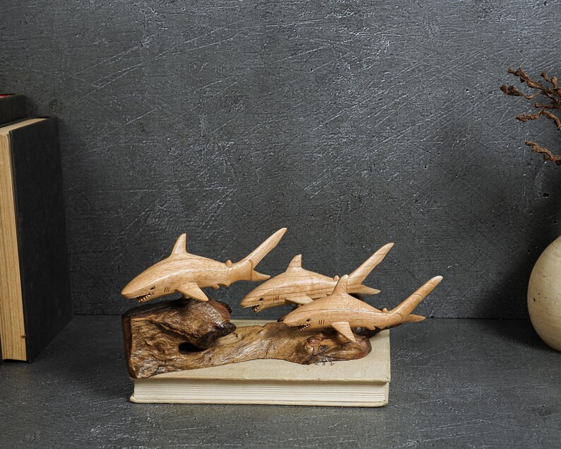 May include: A wooden sculpture featuring three carved sharks attached to a piece of driftwood. The sharks are light brown with visible wood grain, and the base is a dark, textured piece of natural wood resting on a rectangular book.