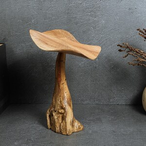 May include: A carved wooden sculpture shaped like a mushroom with a wide, textured cap and a thick, organic-looking stem. The wood displays natural grain patterns and light brown tones, standing upright as a singular decorative object.