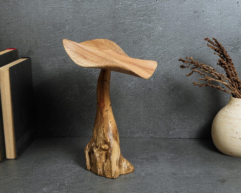 May include: A carved wooden sculpture shaped like a mushroom with a wide, textured cap and a thick, organic-looking stem. The wood displays natural grain patterns and light brown tones, standing upright as a singular decorative object.