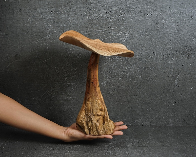 May include: A hand-carved wooden sculpture shaped like a mushroom with a wide, wavy cap and a thick, textured stalk. The wood displays natural light brown and tan grain patterns, with a smooth finish on the cap and a rugged, organic base.