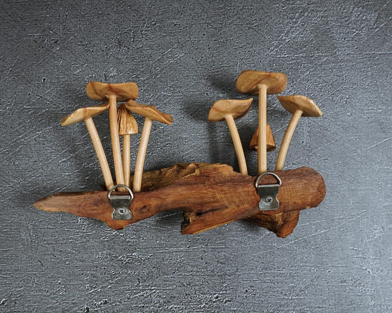 May include: A wall-mounted coat rack crafted from a natural piece of driftwood, featuring two silver-toned metal D-ring hangers. The top of the wood is adorned with several carved wooden mushroom sculptures with light brown caps and stems.