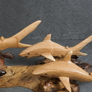 May include: Three light brown wooden shark carvings mounted on a piece of natural driftwood. Each shark figure features carved fins, a tail, and gill slits. The sharks are positioned at different angles, showcasing smooth wood grain textures.