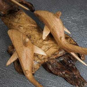 May include: Two carved wooden shark figures mounted on a piece of natural driftwood. The sharks are light brown with visible wood grain, featuring smooth fins and tails. The driftwood base is dark brown with a rough, textured, and organic surface.