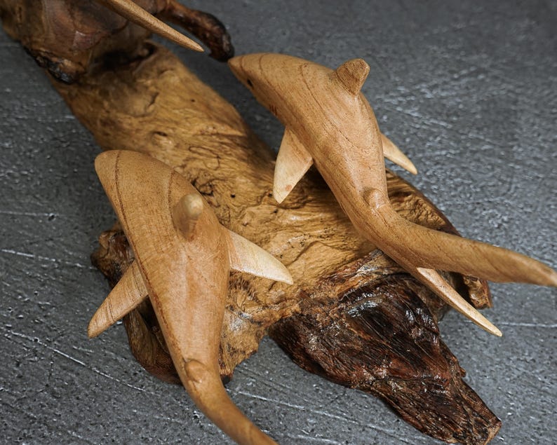 May include: Two carved wooden shark figures mounted on a piece of natural driftwood. The sharks are light brown with visible wood grain, featuring smooth fins and tails. The driftwood base is dark brown with a rough, textured, and organic surface.