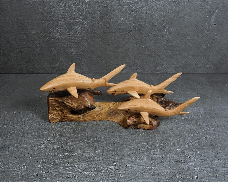 May include: A hand-carved wooden sculpture featuring three sharks attached to a piece of natural driftwood. The sharks are light brown with smooth, polished surfaces, while the base is dark, textured, and irregular in shape.