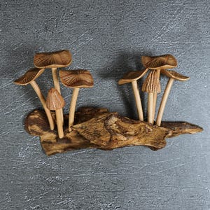 May include: A decorative sculpture featuring seven carved wooden mushrooms with detailed gills under their caps, attached to a piece of textured, natural-looking driftwood. The mushrooms vary in height and cap size, showcasing a light brown wood grain.